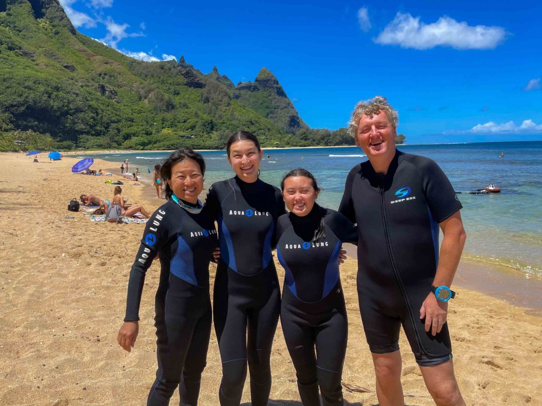 Four divers in blue wetsuits prepping on the sandy beach of Kauai, ready for an underwater adventure.