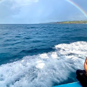 A woman stands on the deck of Fathom Five Divers' boat, a rainbow spanning the sky in the backdrop in Kauai, Hawaii.