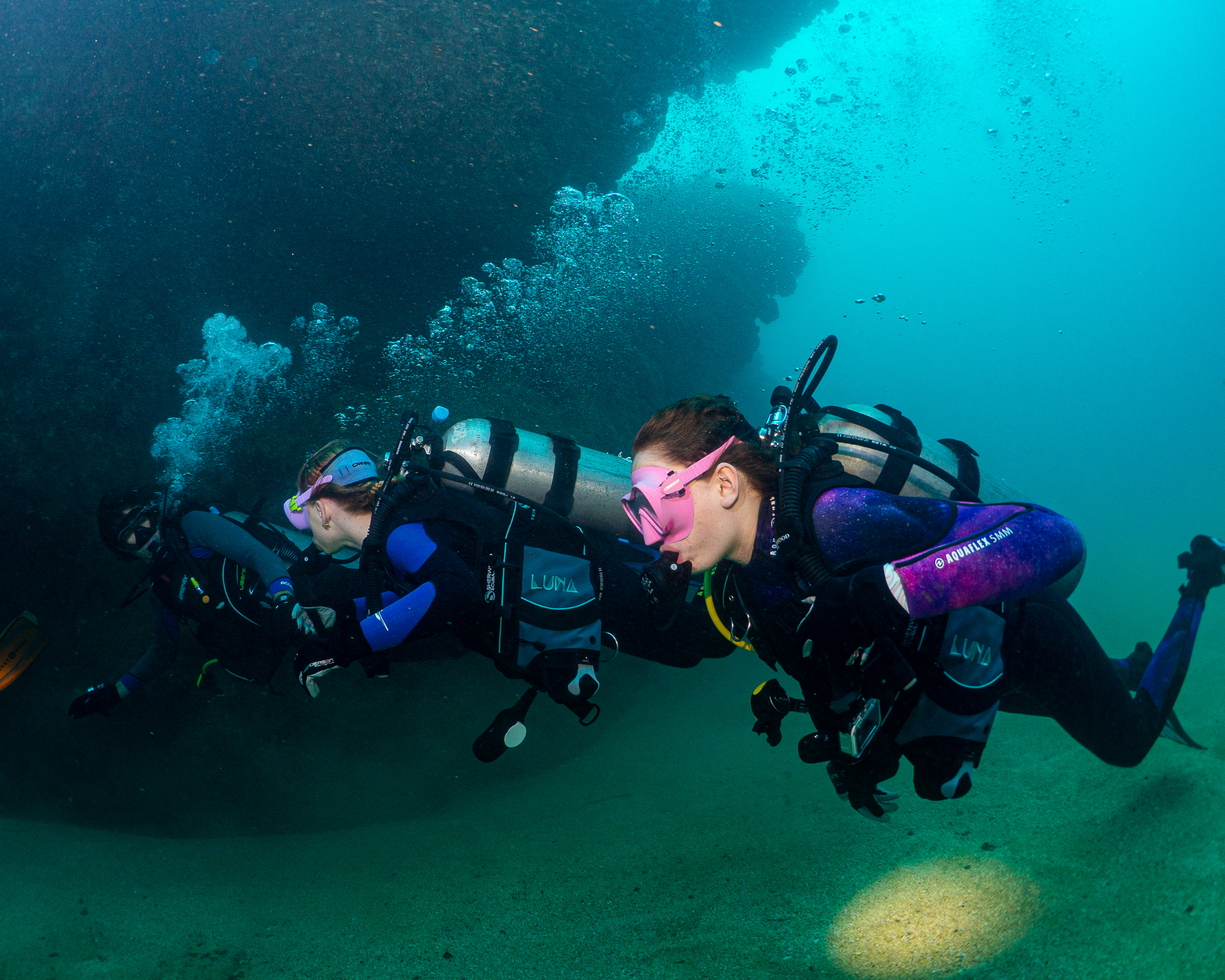 Unity in the Deep: Scuba Divers at Sheraton Caverns, Kauai” A striking image of three scuba divers swimming in unison at the Sheraton Caverns in Kauai, Hawaii, captured in the style of Zeiss Batis 18mm f/2.8, with hues of dark violet and dark cyan