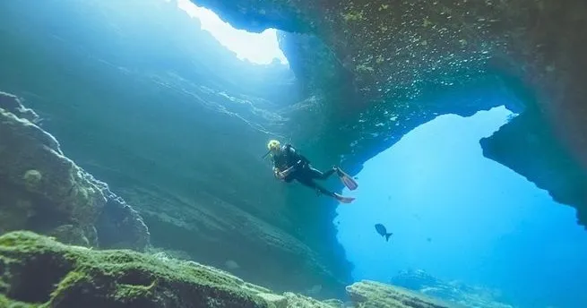 Ethereal Exploration: Diving through Tunnels in Kauai with Fathom Five Divers A diver is swimming through a rocky underwater cave during a dive in Tunnels, Kauai, with Fathom Five Divers