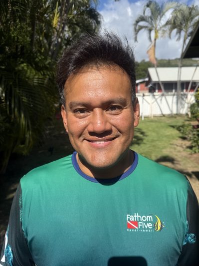 Smiling person in a green 'Fathom Five Kauai-Hawaii' shirt outdoors with palm trees.