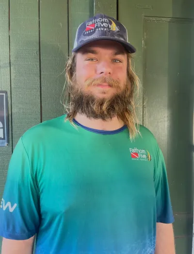Person with a beard wearing a cap and a green Fathom Five shirt, standing against a wooden background.